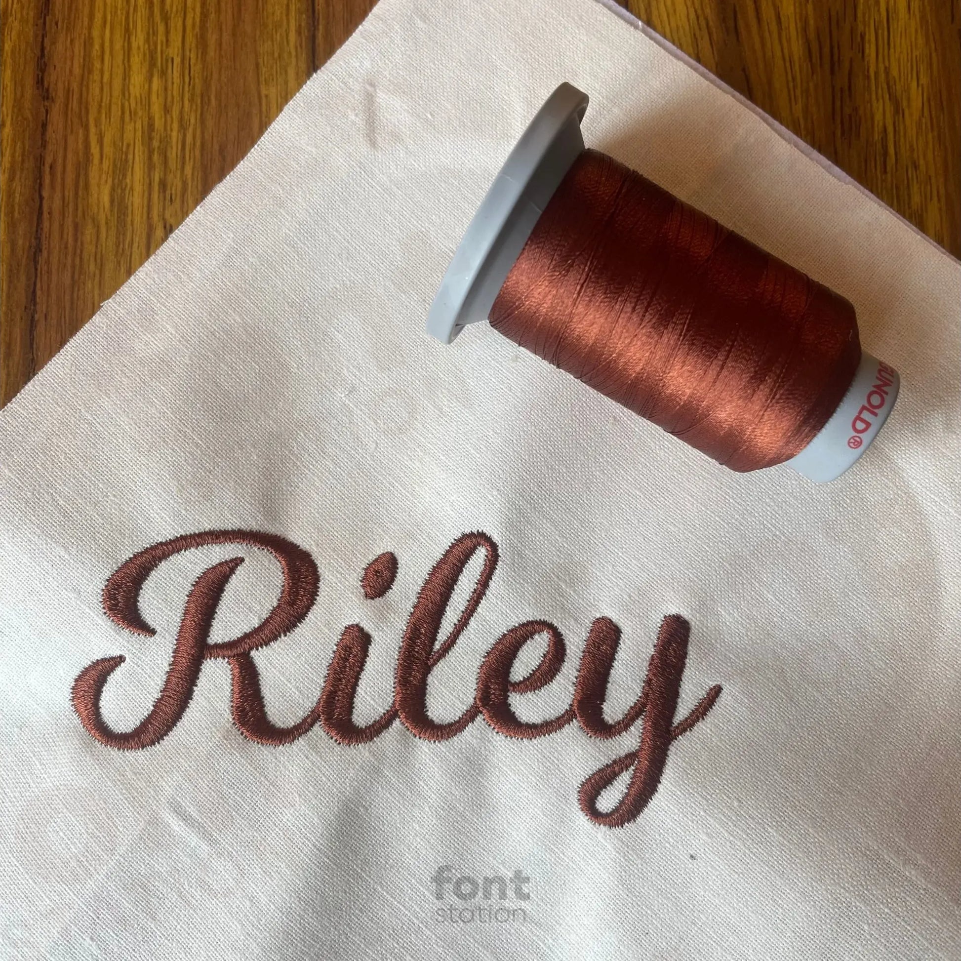 Spool of brown thread on a white fabric with 'Riley' embroidered on it using the Casual Script embroidery font.